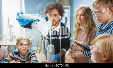 Scuola elementare Scienza Aula: Insegnante entusiasta spiega la chimica a diversi gruppi di bambini, mostra loro come mescolare le sostanze chimiche in Beakers Foto Stock