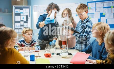 Scuola elementare Scienza Aula: Insegnante entusiasta spiega la chimica a diversi gruppi di bambini, mostra loro come mescolare le sostanze chimiche in Beakers Foto Stock