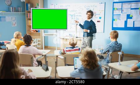 L'insegnante di fisica della scuola elementare usa la lavagna interattiva digitale con il modello di mock-up dello schermo verde. Conduce le lezioni in classe piena di intelligenza Foto Stock