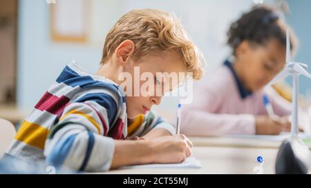 In classe scuola elementare Brilliant caucasic Boy scrive in Eserctaccuino, prendendo prova e scrivendo esame. Classe Junior con gruppo di Foto Stock