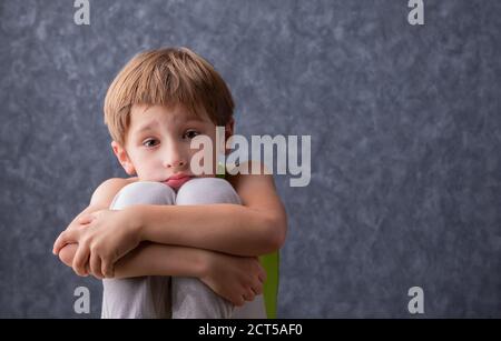 Un bambino triste si siede abbracciando le ginocchia. Un ragazzo di età elementare è triste. Scolaro disunì su uno sfondo grigio. Foto Stock