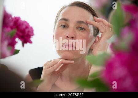 Una donna di mezza età esamina le rughe su uno sfondo di fiori. Cura per la pelle invecchiata. Foto Stock