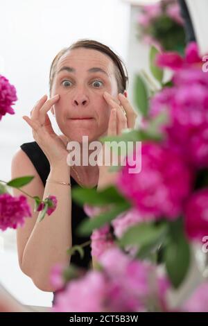 Una donna di mezza età esamina le rughe su uno sfondo di fiori. Cura per la pelle invecchiata. Foto Stock