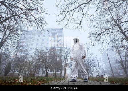 Intera lunghezza di ambientalista maschile in tuta radiante in piedi sul marciapiede su strada foggy. Ecologo con uniforme protettiva bianca e maschera a gas. Concetto di ecologia e inquinamento ambientale. Foto Stock