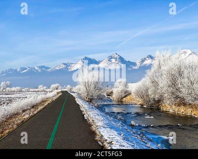 Paesaggio invernale di pista ciclabile accanto al fiume in congelato campo e montagne innevate sullo sfondo Foto Stock