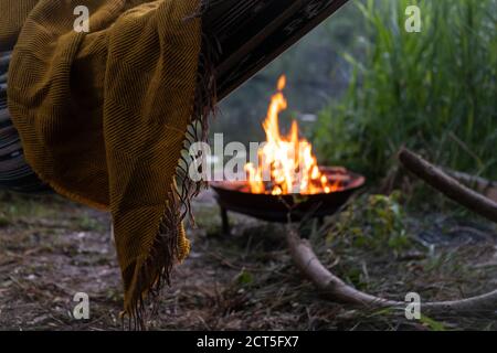 Coperta di senape sull'amaca boho accanto al lago e al fuoco in prefit al crepuscolo Foto Stock