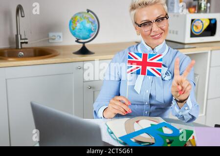 cool insegnante di inglese moderno tenere una lezione online tramite webcam sul computer portatile, guarda lo schermo sorridente, gran bretagna bandiera in mani Foto Stock