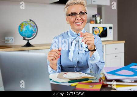 elegante bionda insegnante caucasica femminile avendo classe online via video chat su pc portatile, si siede a tavola a casa in interni moderni, sorridendo gioiosamente, e Foto Stock