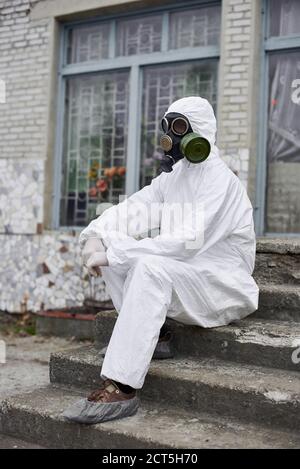 Ambientalista maschile in maschera a gas seduto sui gradini di una vecchia costruzione. Ricercatore che indossa uniforme protettiva bianca, copriscarpe e guanti. Concetto di ecologia e inquinamento ambientale. Foto Stock