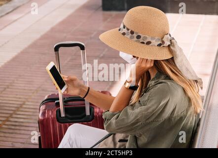 Vista laterale di donna irriconoscibile in cappello e maschera seduta su panca vicino valigia e smartphone di navigazione sulla stazione ferroviaria durante la pandemia Foto Stock