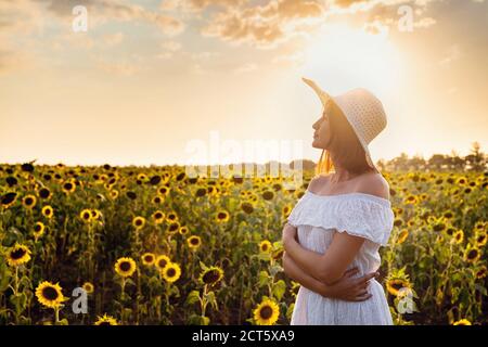Bella giovane donna che gode la natura sul campo di girasoli al tramonto. Donna asiatica in un vestito bianco carino e cappello gode estate e vacanza. Foto Stock