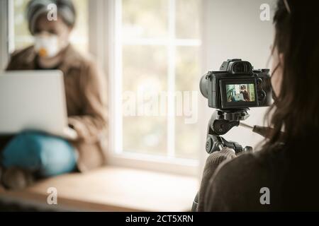 Backstage delle riprese. Messa a fuoco selettiva sulla fotocamera digitale in primo piano. Donna in maschera di faccia con il computer portatile seduto vicino alla finestra su sfondo sfocato Foto Stock