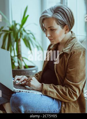 Elegante donna matura lavora portatile seduta sul davanzale della finestra. Vista laterale della moderna Lady dai capelli d'argento con digitazione casual sulla tastiera del computer Foto Stock