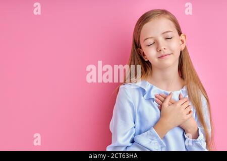 misericordiosa bambina caucasica esprime grazie alla macchina fotografica, bambina con capelli lunghi in posa isolata su sfondo rosa Foto Stock