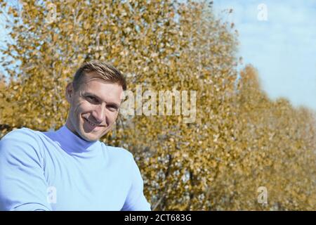 ritratto di un sportivo di mezza età sorridente e guardando lo spettatore. attività autunnale all'aperto. uomo caucasico riposarsi dopo l'allenamento su uno sfondo di alberi autunnali. stile di vita attivo all'aria aperta. Foto Stock