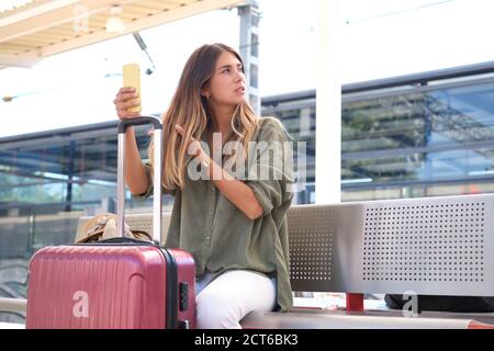 Giovane donna che usa il suo smartphone, pettinando i capelli con le dita e appoggiandosi sulla valigia, in attesa del treno in una stazione ferroviaria. Travele Foto Stock
