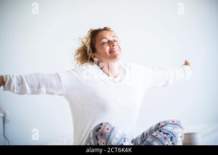 Felice bella donna svegliarsi la mattina a casa o camera d'albergo godendo la vita con un sorriso e. apertura delle braccia per allungare e iniziare la nuova giornata - Foto Stock