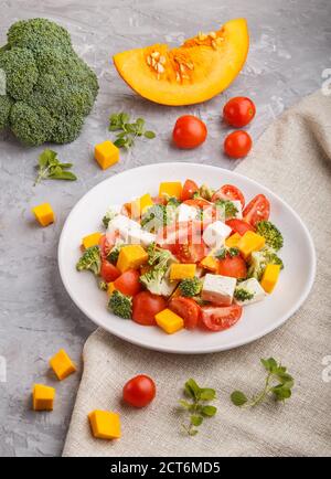 Insalata vegetariana con broccoli, pomodori, formaggio feta e zucca su piastra bianca in ceramica su fondo grigio in cemento e tessuto di lino, vista laterale, Foto Stock