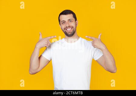 Giovane uomo con barba in una t-shirt bianca faccina sorridente che mostra e punta con le dita denti e bocca. Concetto di salute. Si trova su sfondo giallo isolato. Foto Stock