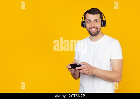 Giovane uomo con barba in maglietta bianca gamer che gioca a videogiochi con le cuffie sorridenti e guardando la fotocamera. Si trova su uno sfondo giallo isolato. Foto Stock