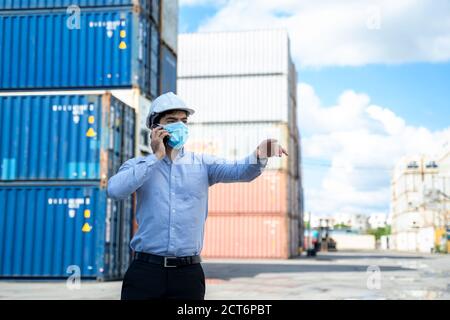 Uomo d'affari che indossa una maschera protettiva per proteggere contro il dipendente Covid-19 casella contenitore di controllo con l'uso di smartphone nel cantiere di spedizione, controllo carico Foto Stock