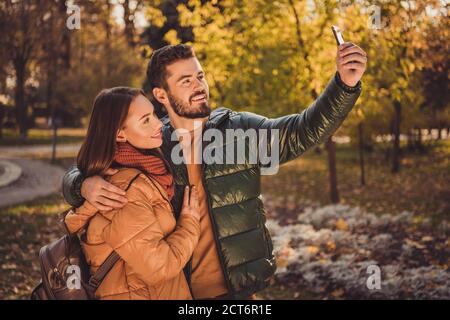 Foto di due persone studenti ragazzo abbraccio ragazza fare selfie su smartphone in autunno città foresta indossare cappotti zaino giallo Foto Stock