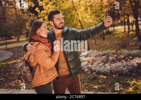 Foto di positivo due persone studenti ragazzo abbracciare ragazza fare selfie su smartphone in autunno parco forestale indossare cappotti zaino Foto Stock