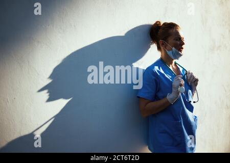 covid-19 pandemic. stanco medico donna moderna in scrub con stetoscopio, maschera medica e guanti di gomma fuori vicino parete. Foto Stock