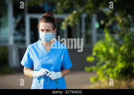 pandemia del coronavirus. donna moderna del medico triste in scrub con stetoscopio e maschera medica all'aperto vicino alla clinica. Foto Stock