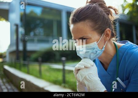 pandemia di coronavirus. infelice medico donna moderna in scrub con stetoscopio e maschera medica seduta fuori vicino clinica. Foto Stock