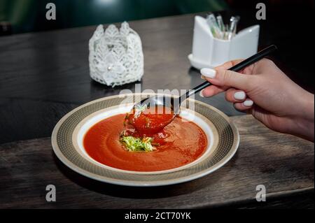 Gazpacho dieta zuppa di pomodoro con erbe e cetriolo su un piatto. Una mano di donna con un cucchiaio di zuppa. Primo piano Foto Stock