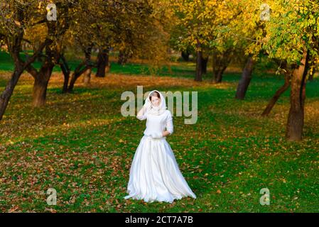 Carina ragazza in lungo vestito bianco di nozze che posa in un percorso rurale tra alberi autunnali nella foresta in atmosfera d'ora d'oro. Foto Stock