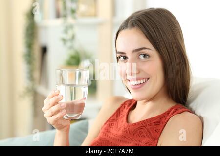 La donna felice sta tenendo un vetro di acqua minerale seduto sopra un divano nel soggiorno di casa Foto Stock
