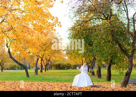 Carina ragazza in lungo vestito bianco di nozze che posa in un percorso rurale tra alberi autunnali nella foresta in atmosfera d'ora d'oro. Foto Stock