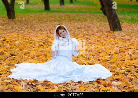 Carina ragazza in lungo vestito bianco di nozze che posa in un percorso rurale tra alberi autunnali nella foresta in atmosfera d'ora d'oro. Foto Stock