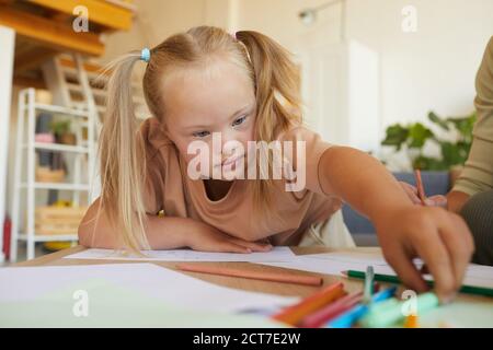 Ritratto della ragazza bionda carina con la sindrome giù disegno e raggiungere per matite mentre godendo la classe di sviluppo, spazio di copia Foto Stock