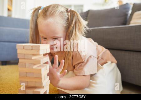 Ritratto di cute ragazza bionda con la sindrome giù giocando giochi da tavolo impilando blocchi di legno mentre si siede sul pavimento a casa, copia spazio Foto Stock