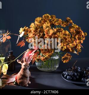 Un bouquet di elio arancio con foglie di uva selvatica in un vaso di vetro scanalato contro una parete blu scuro. Figurina di porcellana di lepre. Foto Stock