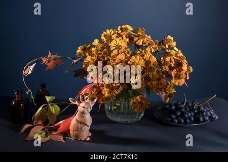 Un bouquet di elio arancio con foglie di uva selvatica in un vaso di vetro scanalato contro una parete blu scuro. Figurina di porcellana di lepre. Foto Stock