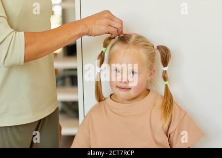 Primo piano di madre premurosa irriconoscibile che combatte i capelli della ragazza carina con la sindrome di Down e legandola in pigtail, spazio di copia Foto Stock