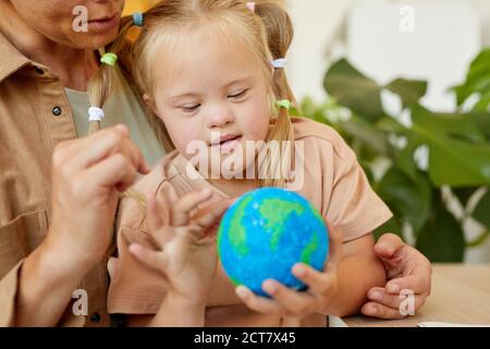 Primo piano ritratto di cute ragazza con sindrome di Down tenendo il modello di pianeta mentre studiando a casa con madre, spazio di copia Foto Stock