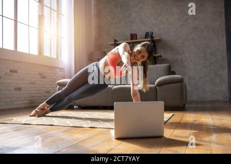 La giovane donna segue con un computer portatile una palestra esercizi. È a casa a causa della quarantena di coronavirus codiv-19 Foto Stock