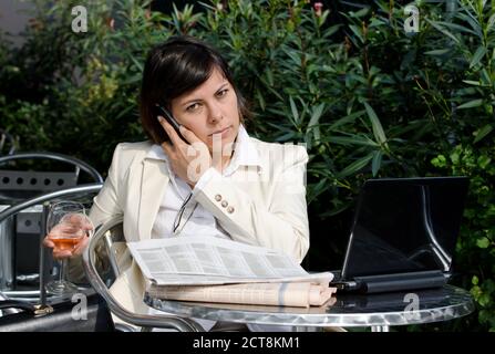 Donna di affari seduto con il suo Personal computer e giornale finanziario e utilizzando Smart Phone su un tavolo esterno. Foto Stock