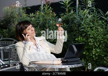 Donna di affari seduto con il suo Personal computer e giornale finanziario e utilizzando Smart Phone su un tavolo esterno. Foto Stock