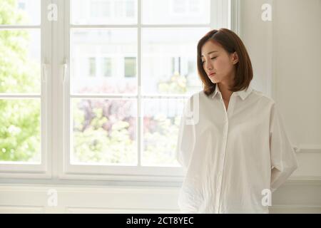 giovane donna asiatica in piedi dalla finestra a casa guardando annoiata e triste Foto Stock