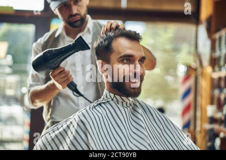 Gioioso uomo dai capelli scuri che si asciuga i capelli in un salone Foto Stock