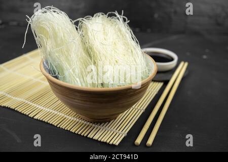 noodle di riso di vetro in una ciotola, fondo asiatico della cucina con stuoia di bambù, chopsticks, salsa di soia su sfondo nero Foto Stock