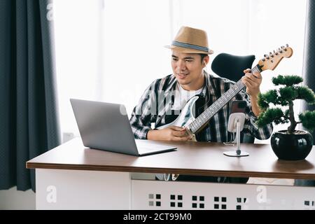 Uomo asiatico che indossa le cuffie che suona la chitarra online sul computer portatile e guarda lo smartphone dal vivo sui social media a casa. Rimani a casa. Quarantena. Foto Stock