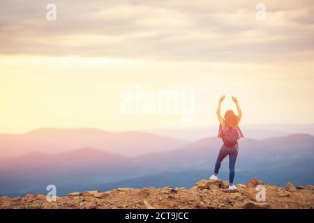 Felice ragazza si trova sulla cima della montagna, solleva le mani sullo sfondo del tramonto, fotografia dal retro Foto Stock