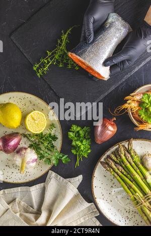 Le mani in guanti neri sostengono il pesce trota su asse di taglio di pietra nera circondato erbe, cipolla, aglio, asparagi, gamberi, gamberi in ceramica piastra Nero Foto Stock